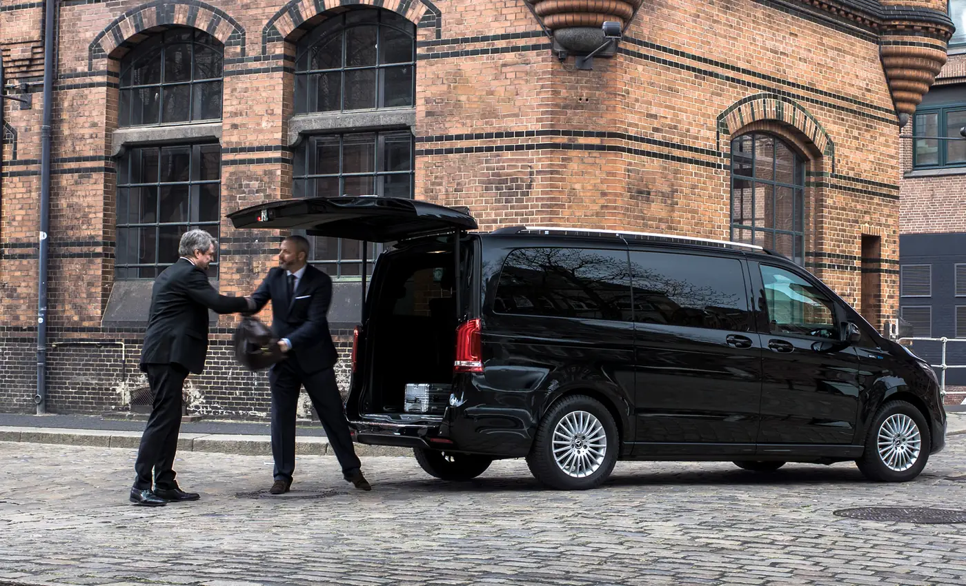 A chauffeur stows a suitcase in the back of a black Mercedes-Benz van