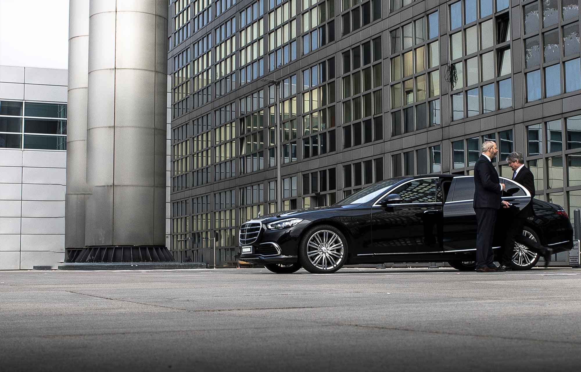 A chauffeur opens the rear door of a black sedan for his passenger in front of an office facade