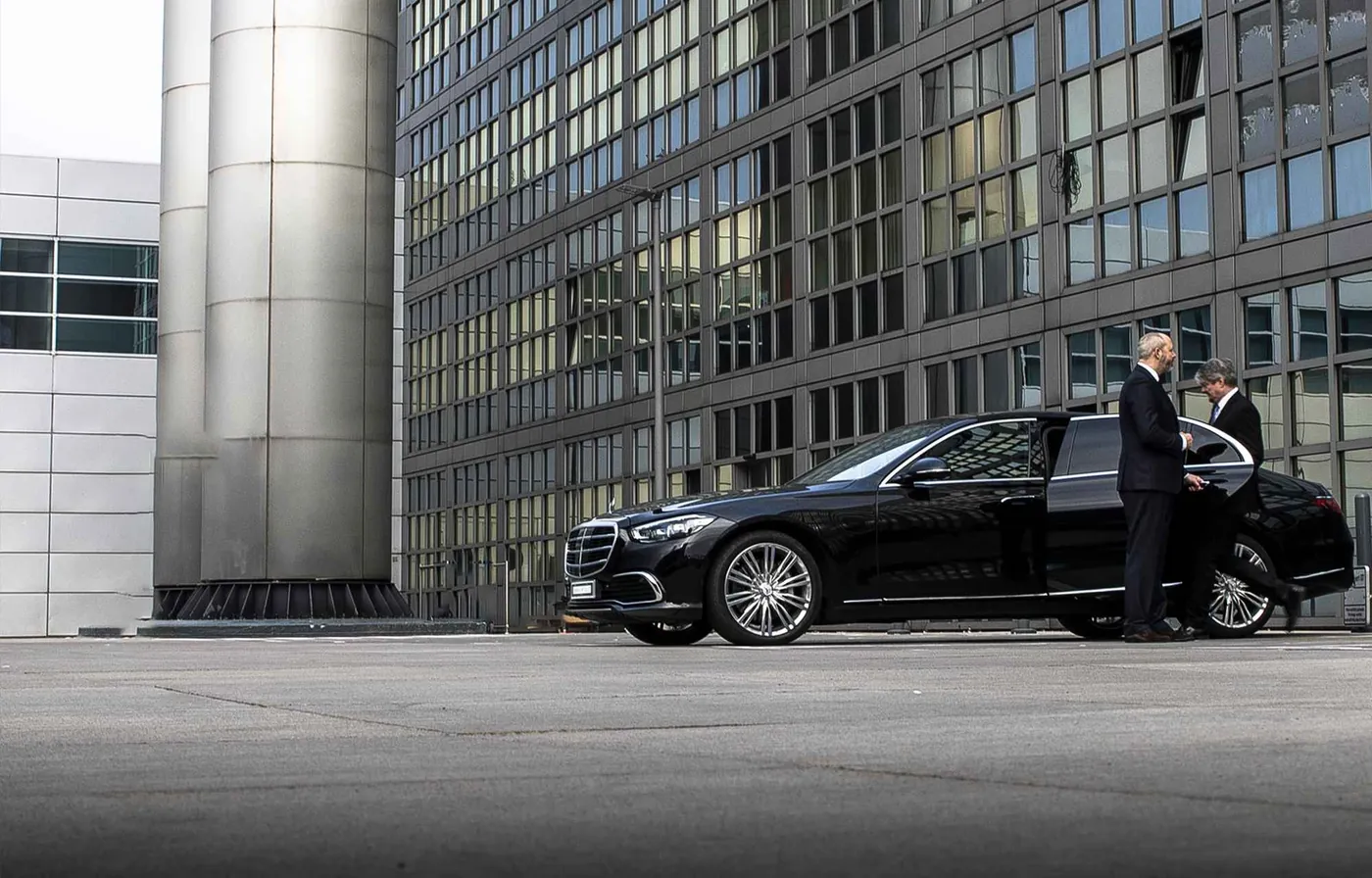Chauffeur im Anzug wartet an der geöffneten Fondtür einer schwarzen Limousine vor einem Bürogebäude