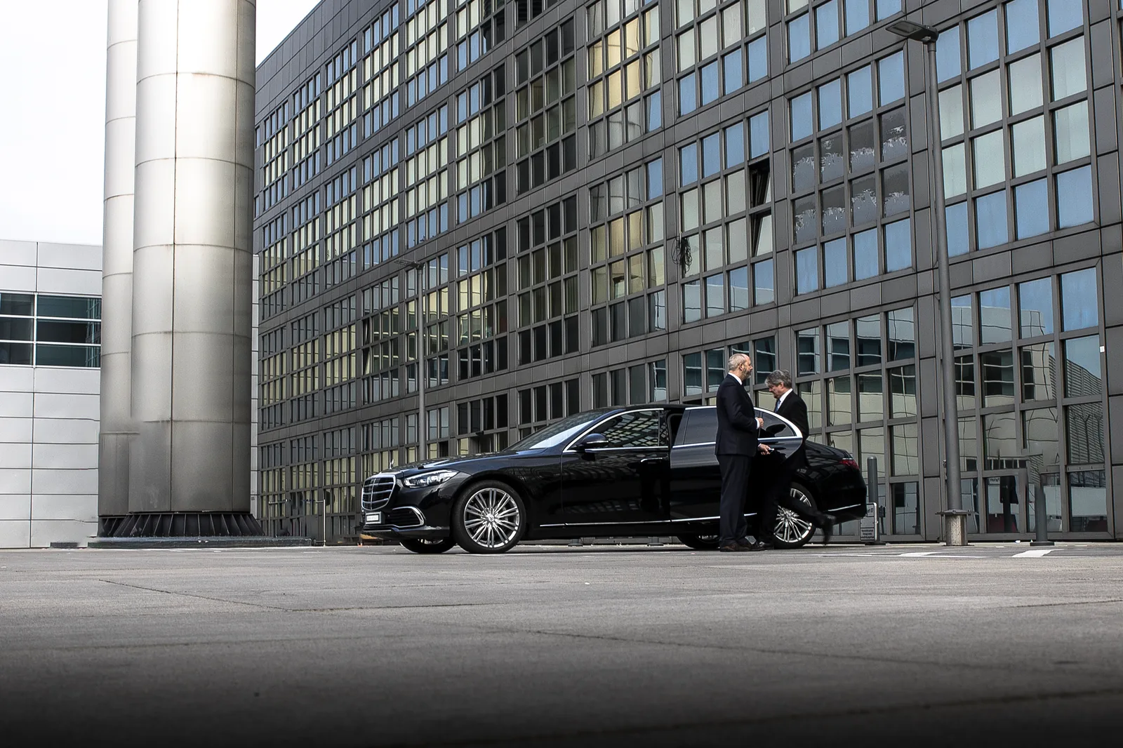 Chauffeur im Anzug an der geöffneten Fondtür einer schwarzen Mercedes-Benz Limousine vor einem Bürogebäude