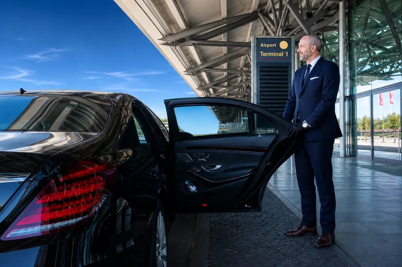 Chauffeur im Anzug hält die Fondtür einer schwarzen Limousine vor dem Eingang zu Terminal 1 auf