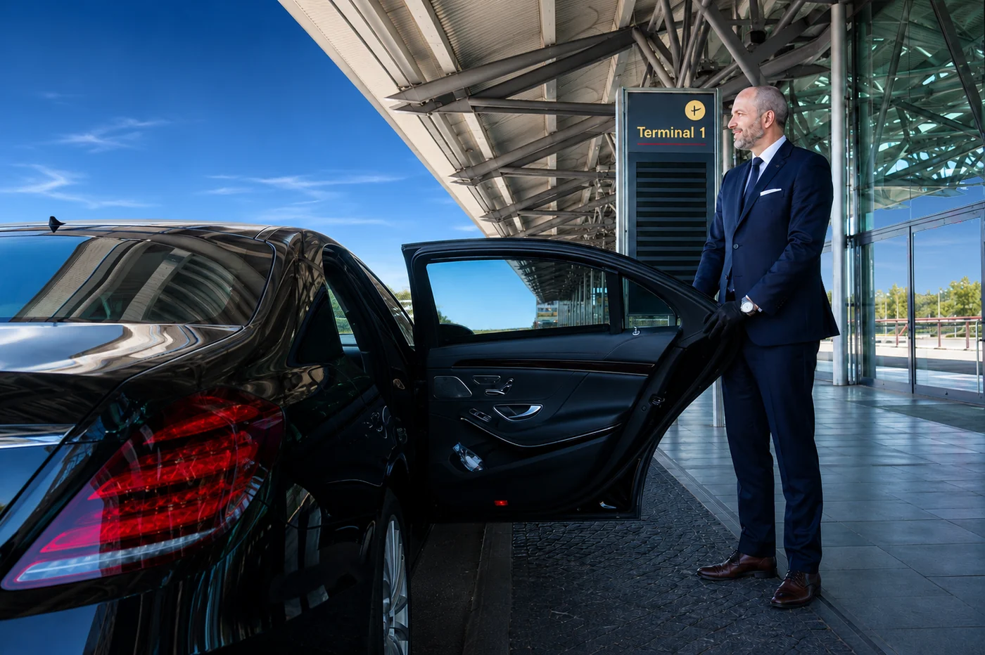 A chauffeur in a suit opens the rear door of a black sedan in front of an airport terminal